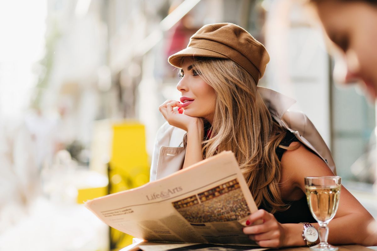 Dreamy woman in brown cap thinking about something, propping face with hand and holding newspaper
