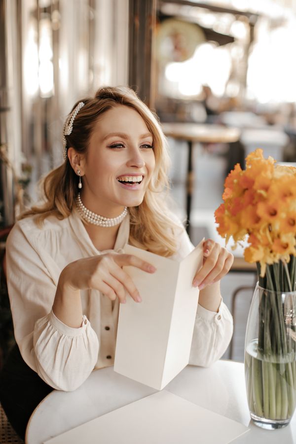 Joyful curly blonde lady in stylish white blouse, pearl necklace smiles sincerely, sits in street c