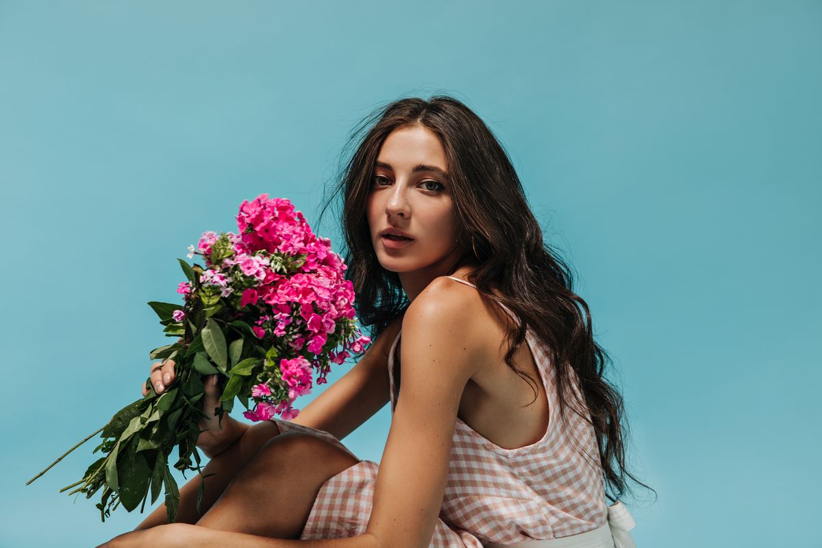 Wonderful girl with wavy hair in modern plaid light clothes holding pink flowers and looking into c